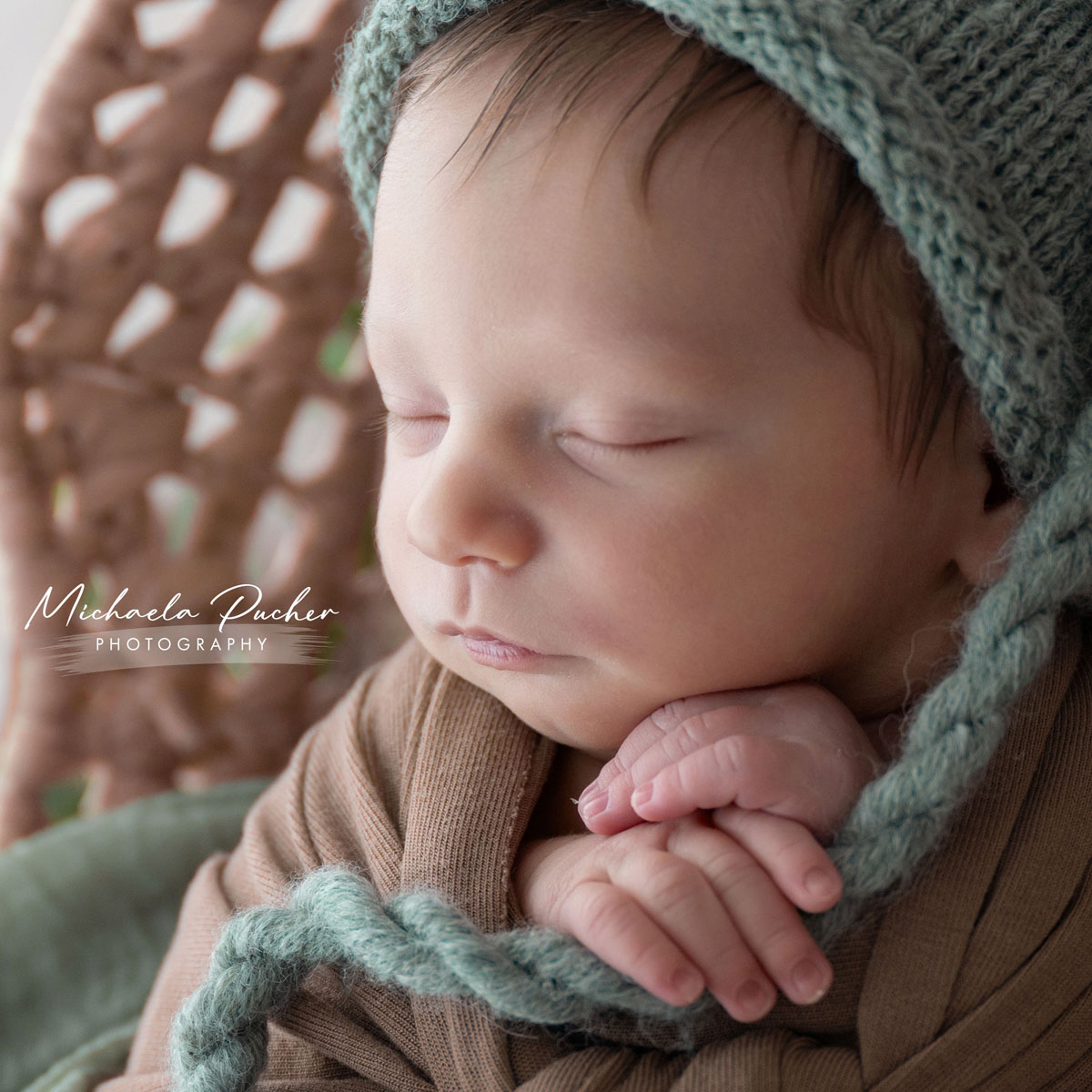 Nahaufnahme eines schlafenden Neugeborenen, eingewickelt in braune Tücher und mit einer grünen Strickmütze, die kleinen Hände unter dem Kinn.